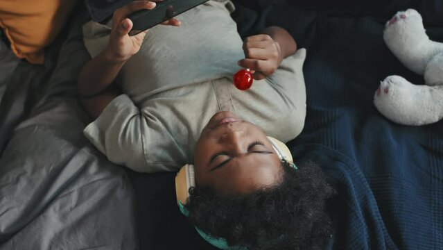 Top view medium of ten-year-old Black boy wearing over-ear headphones, lying on bed with legs up on wall in his room at daytime, having lollipop and using smartphone