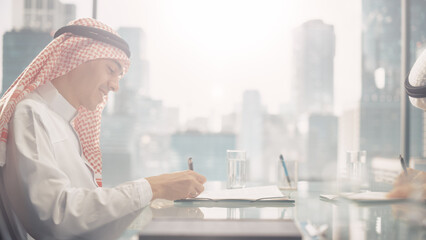 Two Successful Emirati Businessmen in White Traditional Outfit Sitting in Office and Signing Contract. Business Partners Sing Lucrative Investment. Saudi, Emirati, Arab Businessman Concept. - obrazy, fototapety, plakaty