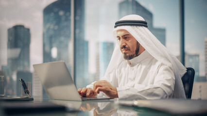 Successful Arab Businessman in White Traditional Kandura Sitting in Office and Working on Laptop Computer. Business Manager Planning Investments. Saudi, Emirati, Arab Businessman Concept.