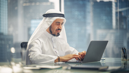 Arab Businessman in White Traditional Outfit Sitting in Office and Working on Laptop Computer. Business Manager Make Successful Investment Deal. Saudi, Emirati, Arab Businessman Concept.