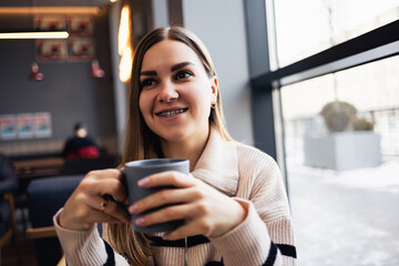 Beautiful smiling calm young woman drinking coffee looking out the window while sitting at a table in a restaurant. Morning coffee