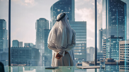 Successful Muslim Businessman in Traditional White Kandura Standing in His Modern Office Looking out of the Window on Big City with Skyscrapers. Successful Saudi, Emirati, Arab Businessman Concept.