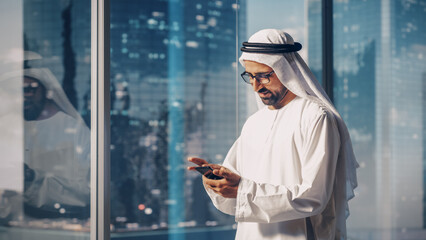 Successful Muslim Businessman in Traditional White Outfit Standing in His Modern Office, Using Smartphone Next to Window with Skyscrapers. Successful Saudi, Emirati, Arab Businessman Concept. - obrazy, fototapety, plakaty