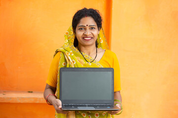 Portrait of indian woman wearing sari showing empty laptop screen. Female wearing traditional cloths holding blank computer display. mockup. advertisement, against orange background,