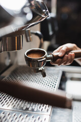 Cropped view of barista holding portafilter with coffee near blurred coffee machine on foreground in cafe.