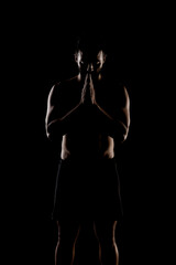 Side lit muscular Caucasian man silhouette. Athlete praying against black background.
