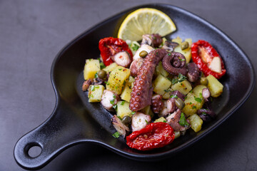 Warm salad with octopus, potatoes, sun-dried tomatoes, capers and lemon in a black small pan. Close-up, black background.