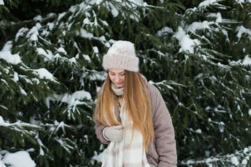 Happy woman in winter forest during snowfall, enjoying winter. Millennial young woman with long hair in beige warm outfit walking in the snowy park in nature near fir trees, being careless and free.