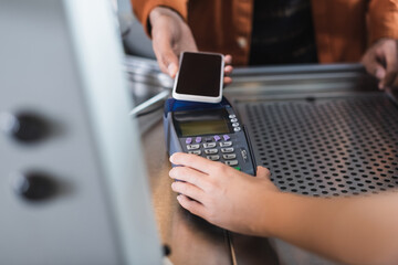 Cropped view of barista holding payment terminal near african american customer with cellphone in cafe.