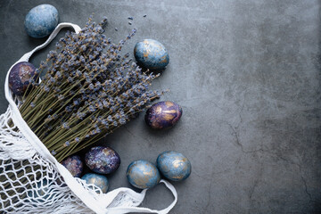 Easter card with a copy of the place for the text. Purple, blue and golden eggs with a bouquet of lavender in a string bag on a dark background. The purple hue trend of 2022 is very peri. 