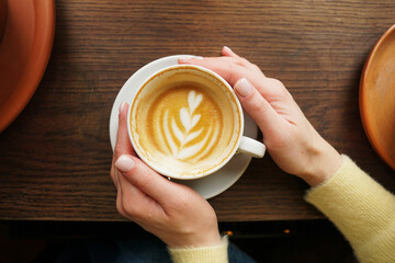Woman hands in yellow sweater holding cup of fresh coffee with latte art on wooden background. Cappuccino closeup. Coffee break. Hygge concept.