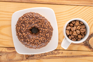 One sweet chocolate donut on a white saucer with a cup of coffee beans, on a wooden table, macro, top view.
