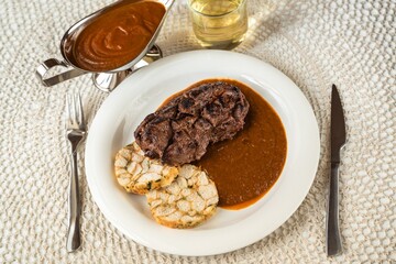 Deer meat, plum sauce and dumpling on plate, cutlery, sauce in bowl, wine in glass on table.