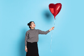 Nice young smiling woman with natural make up standing with heart balloon isolated on blue background. Valentines Day concept