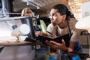 African american barista writing on notebook near coffee machine and payment terminals in cafe.