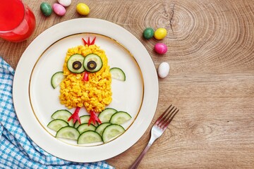 Scrambled egg easter chick made it from scrambled eggs,cucumbers,black olives and red hot peppers on plate with wooden background.Art food idea for kids Easter's breakfast.Top view.Copy space