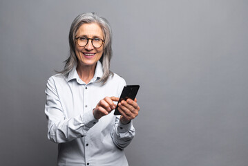 Fototapeta premium Happy business woman in formal shirt touching to the smartphone screen while feeling great with new phone features. Indoor studio shot isolated on grey background