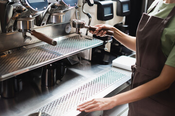 Cropped view of barista in apron holding portafilter near coffee machine in cafe.