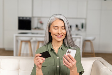 Serene middle-aged grey-haired Asian woman shopping online sitting with credit card and mobile...