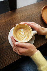 Woman hands in yellow sweater holding cup of coffee with latte art on wooden background. Cappuccino. Coffee break. Hygge concept. Vertical photo