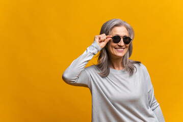 Horizontal shot of retirement female model wears stylish clothes and big sunglasses, posing cheery, buying clothes of last trends against yellow background