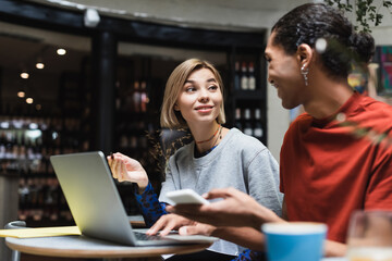 Smiling freelancer talking to african american friend near devices in cafe.