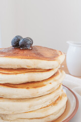Macro close up of stack of pancakes with blueberries on top and maple syrup or honey running down the sides. Pouring jug in background set against a light airy white backdrop.