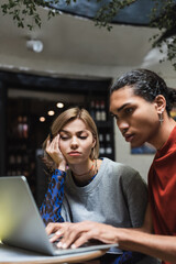 Upset freelancer looking at laptop near african american friend in cafe.