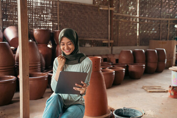 veiled asian woman smiling using tablet while sitting between pottery