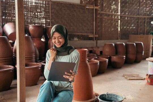 veiled asian woman smiling using tablet while sitting between pottery