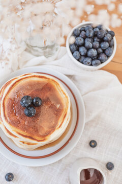 Flat lay image of a stack of pancakes with blueberries on top and maple syrup or honey running down the sides. Fruit, flowers and jug on top a wooden table. Copy space available in light airy backdrop