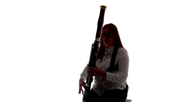 Silhouette woman plays bassoon music sitting on chair in studio, white background, front view. Female musician sits on chair and plays instrument