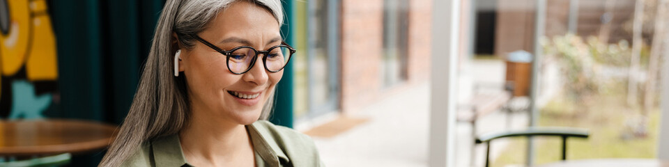 Mature smiling woman using earphone and mobile phone in cafe
