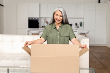 Satisfied stylish senior Asian woman opens parcel carton box, unpacking received gift, feeling...