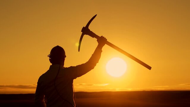 Businessman man in hard hat stands background sunset with pickaxe raised up, back view. Cryptocurrency man stands outdoor with pickaxe in hand and helmet. Mining concept