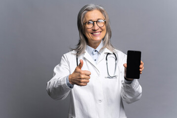 Cheerful senior mature female doctor with a stethoscope in a medical gown posing isolated over gray background, showing a thumbs-up and holding smartphone with empty screen - Powered by Adobe