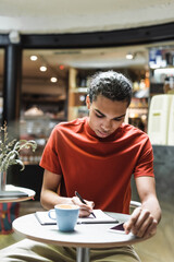 Fototapeta na wymiar African american man writing on notebook near coffee and smartphone in cafe.