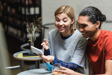 Positive woman pointing at smartphone near african american boyfriend with coffee in cafe.