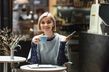 Smiling blonde woman holding cellphone and coffee near notebook in cafe.