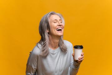 Smiling senior woman in casual wear holding paper cup of coffee isolated over yellow background. Mature happy gray-haired lady standing and laughing, while enjoying coffee to-go