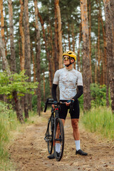 Fototapeta premium Portrait of a male cyclist in a professional outfit standing with a bicycle on a trail in a pine forest on a background of beautiful views and looking away.