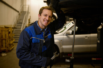 Portrait of a young mechanic in professional uniform, holding a wrench and working in auto service with lifted vehicle, smiles toothy smile lookng at camera. Car repair and maintenance.