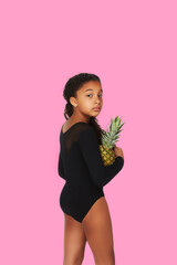 A swarthy girl with black curly hair is half-turned over pink backdrop and holding a pineapple in...