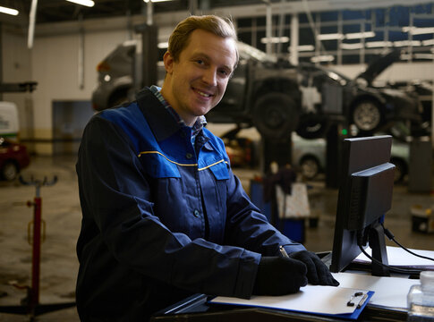 Concentrated auto mechanic technician car engineer smiles looking at camera writing checklist on clipboard and working on monitor while servicing a car in the auto service