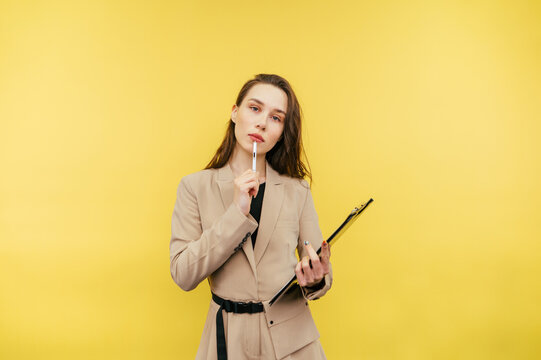 Business woman with pen and tablet stands with a pensive face on a yellow background, looking at the camera and thinking. Isolated