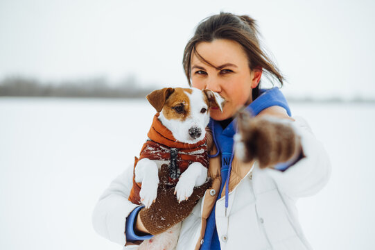 Portrait of a beautiful woman in warm clothes standing with a dog breed Jack Russell in his arms and pointing his finger at the camera on a background of snowy views.