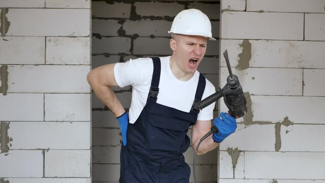 A male construction worker in a helmet with a drill in his hands is messing around and having fun at a construction site