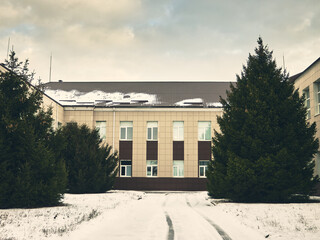 Fototapeta premium Part of a two-story building lined with tiles and a metal tile roof, lush fir trees grow in the courtyard, snow covers the asphalt, winter city landscape