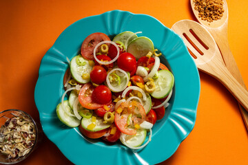 Um prato com salada de vegetais, um copo com especiarias desidratadas e dois talheres de madeira em um fundo laranja.