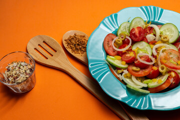 Um prato com salada de vegetais, um copo com especiarias desidratadas e dois talheres de madeira em um fundo laranja.